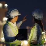 Two_industrial_engineer_using_digital_tablet_for_work_and_discussing_against_the_background_of_electrical_power_plant_at_night_time.
