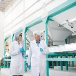 Portrait_of_two_workers_wearing_white_coat_walking_across_clean_production_hall_at_modern_factory,_copy_space